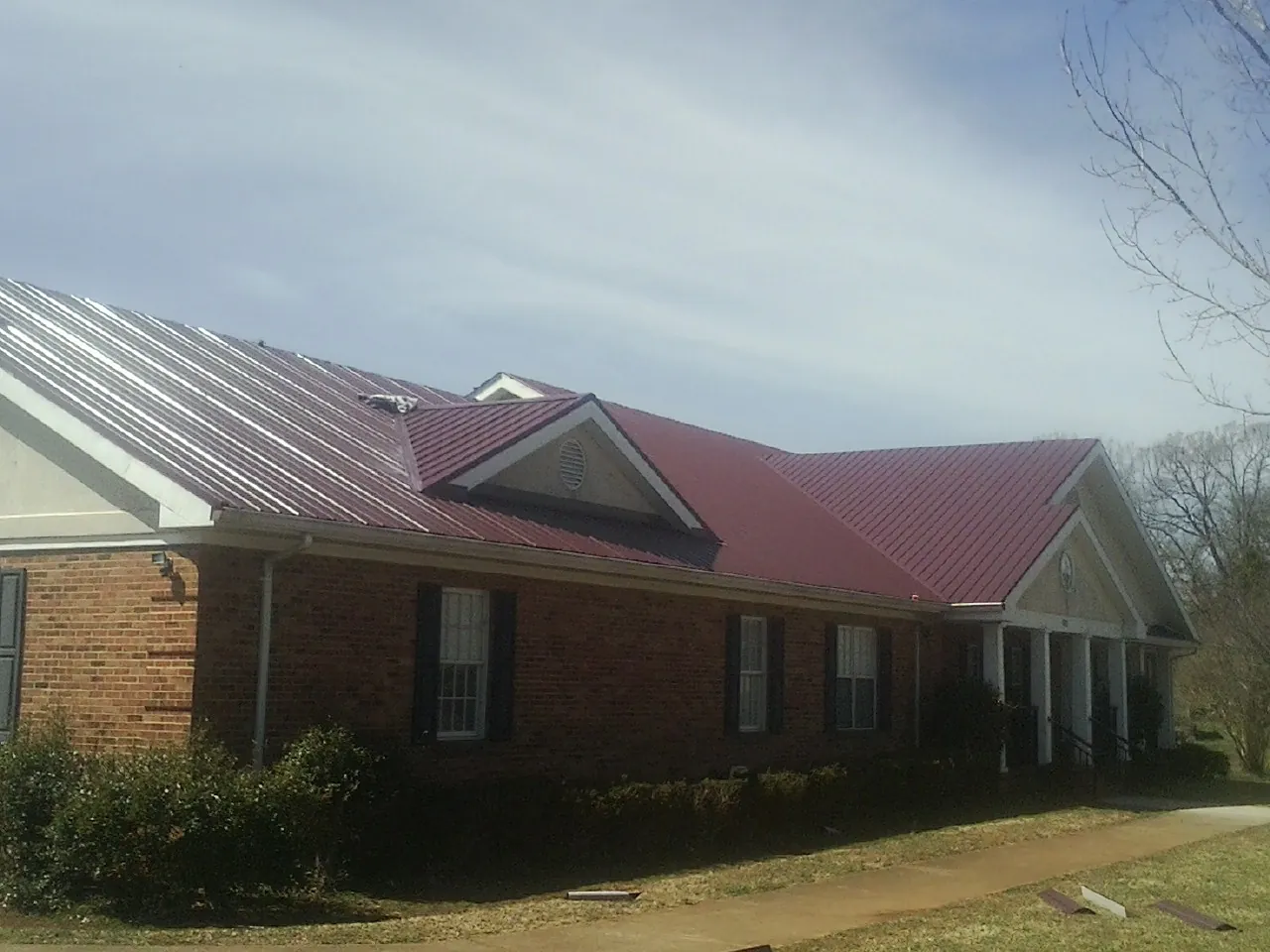 Red metal roof with gabled entries on a long brick building, from a Hail Damage Roof Repair job in Many