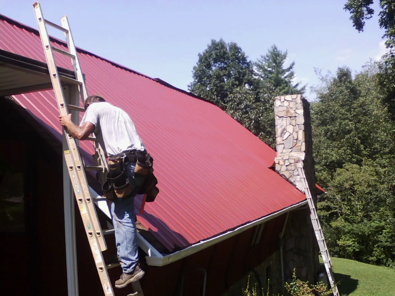 Roofer on a ladder beside a red metal roof and stone chimney — Soffit & Fascia Repair by Roofing Brothers in Many